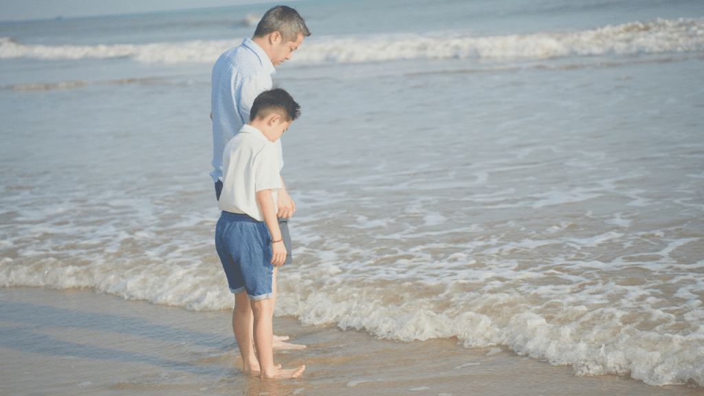 Father and son stand barefoot at the beach as the ocean waves lap their feet.