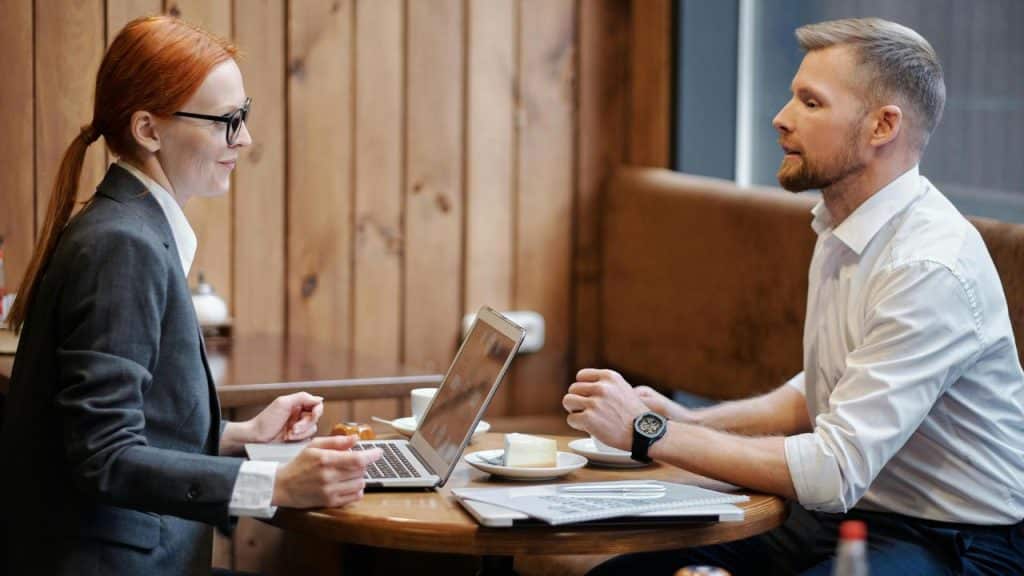 A man and a woman having a business meeting at a café table with a laptop and notebooks.
