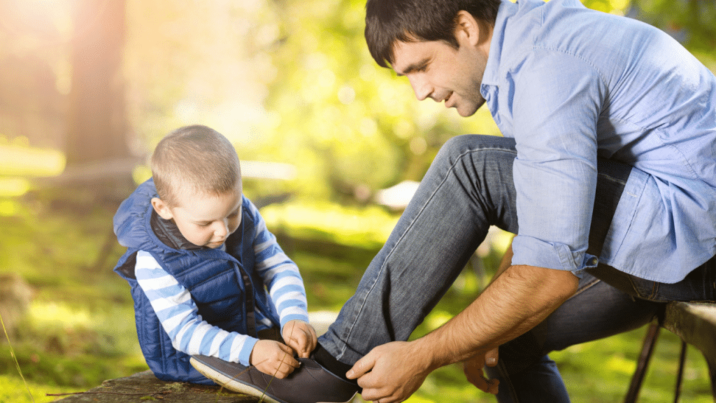 A father and his young son sit outdoors while the boy ties his father's shoe.