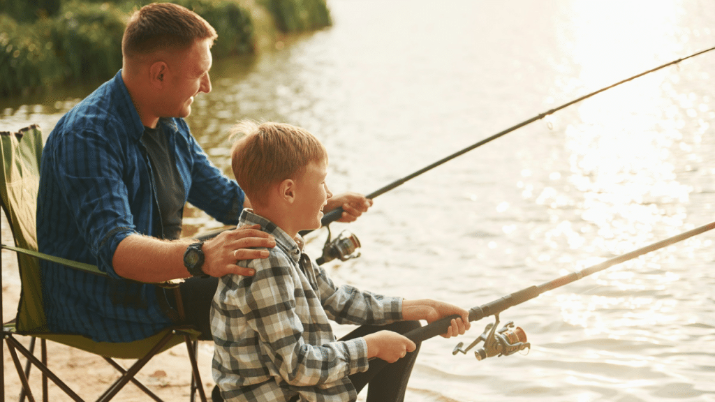 Father and son sitting by the water, smiling, holding fishing rods together.