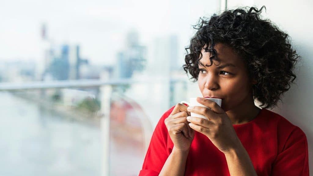 A woman in a red shirt drinking coffee by a window.