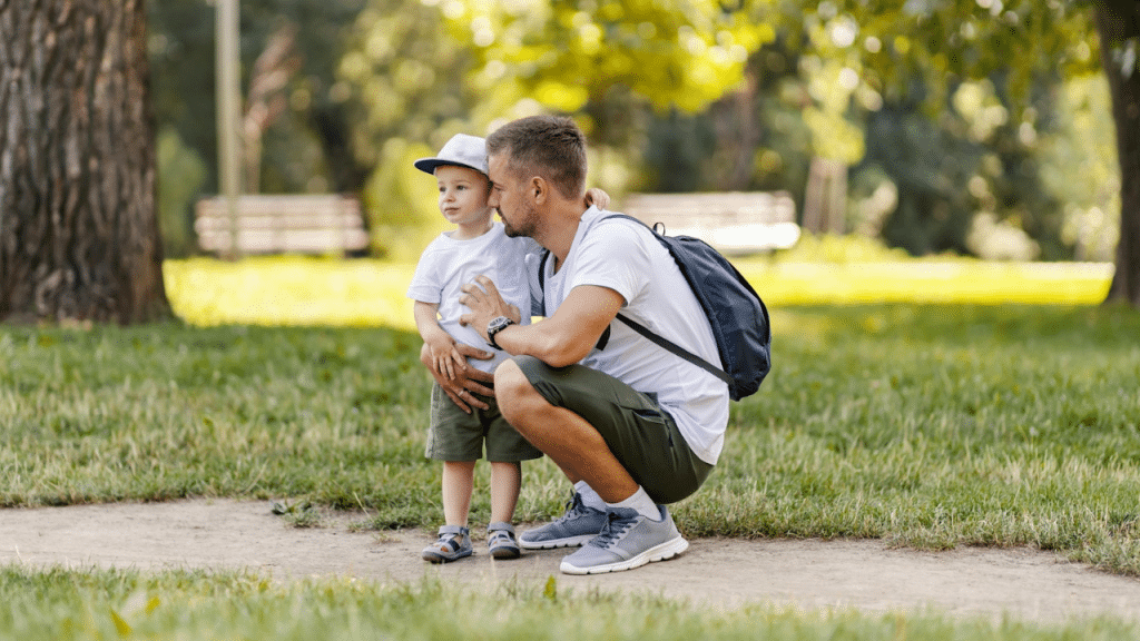 A father kneels down, holding his young son, outdoors in a park setting.