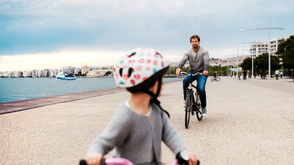 A father and son ride bicycles along a waterfront promenade in a city.