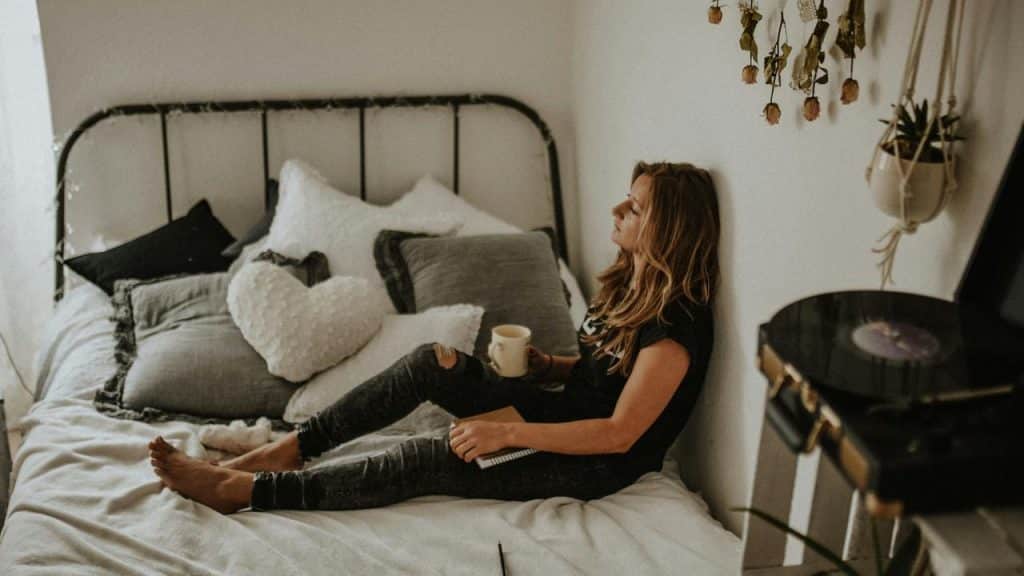 A woman sitting on a bed with a cup of coffee, leaning against the wall.