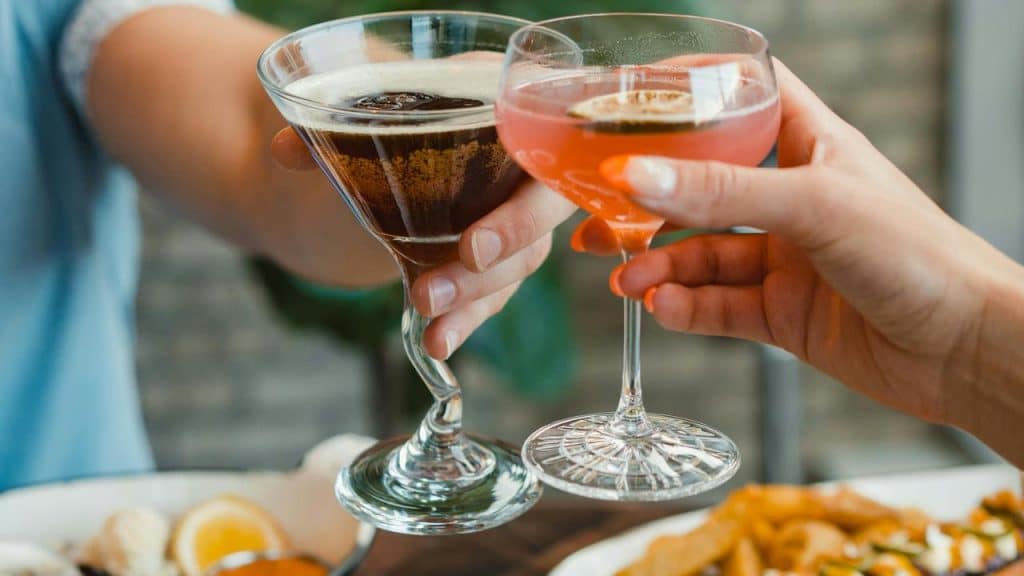 A close-up of two people clinking cocktail glasses over a meal.