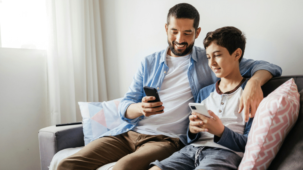 Father and son sit on a couch, smiling, both looking at their smartphones.