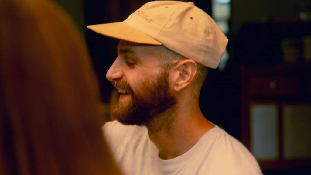 A smiling man wearing a beige cap and a white shirt.