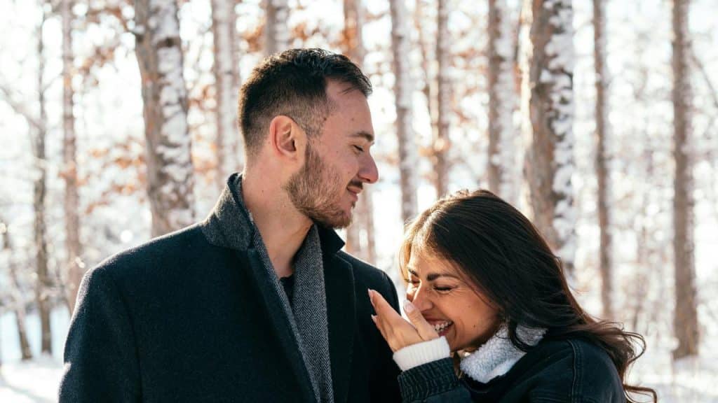 A smiling couple standing close together in a snowy forest.