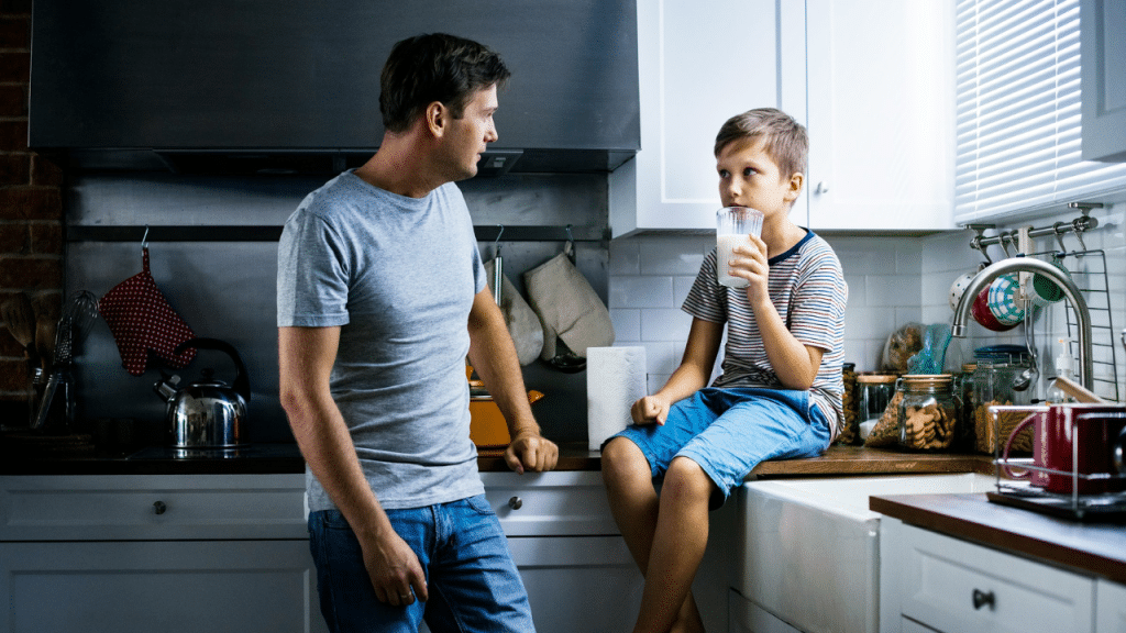 A father and son in a kitchen, the boy sitting on the counter drinking milk.