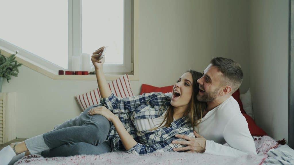 A couple laughing together while taking a selfie on a bed.