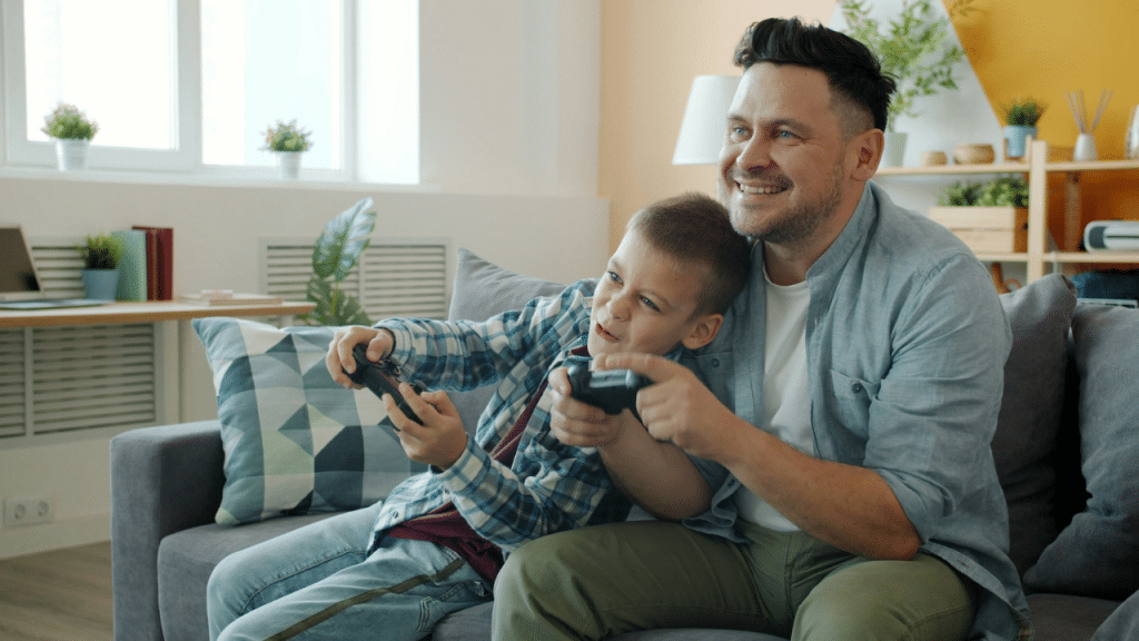 Father and son sit on a couch, smiling, playing video games with controllers.