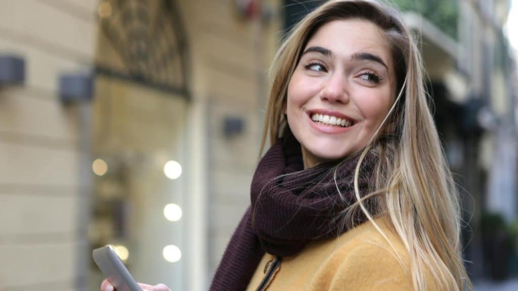 A woman smiling while holding her phone on a city street.