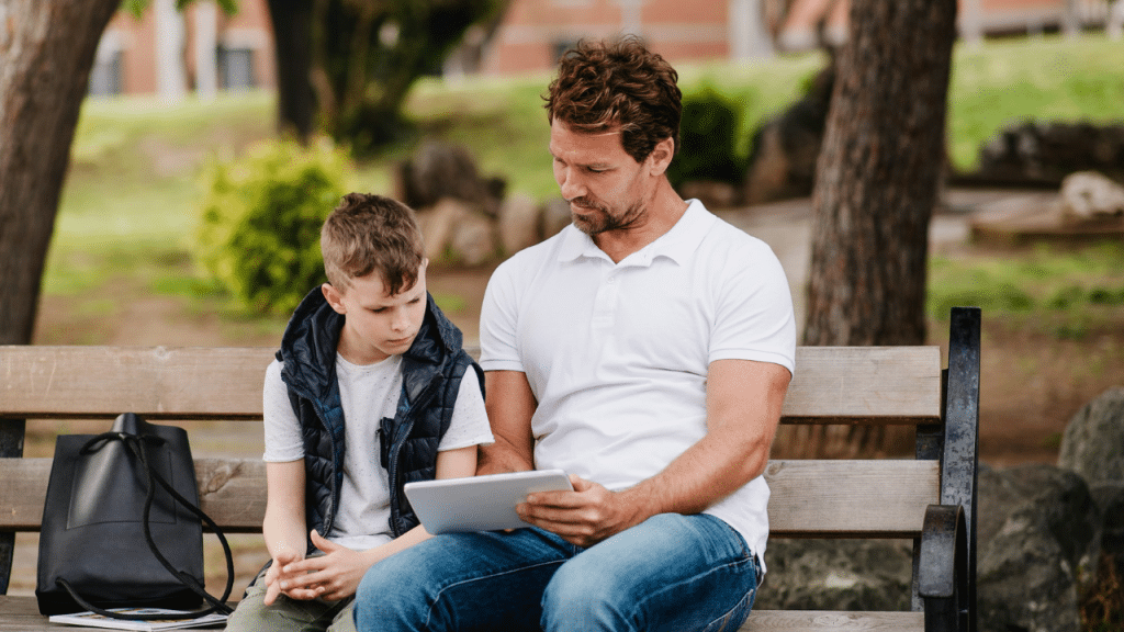 A father and son sit on a park bench, looking at a tablet together.
