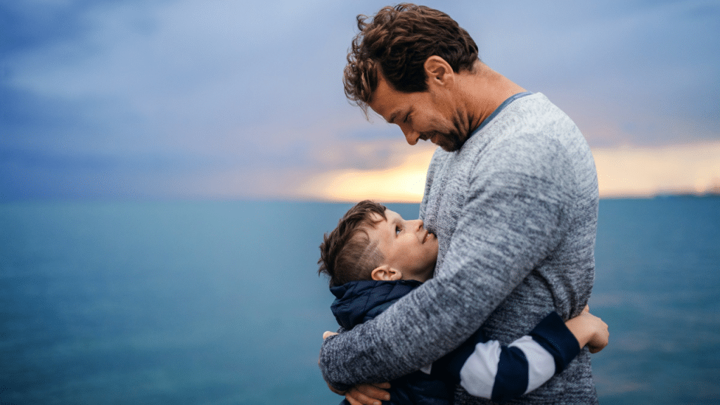 A father and son embrace outdoors near the water under a cloudy sky.