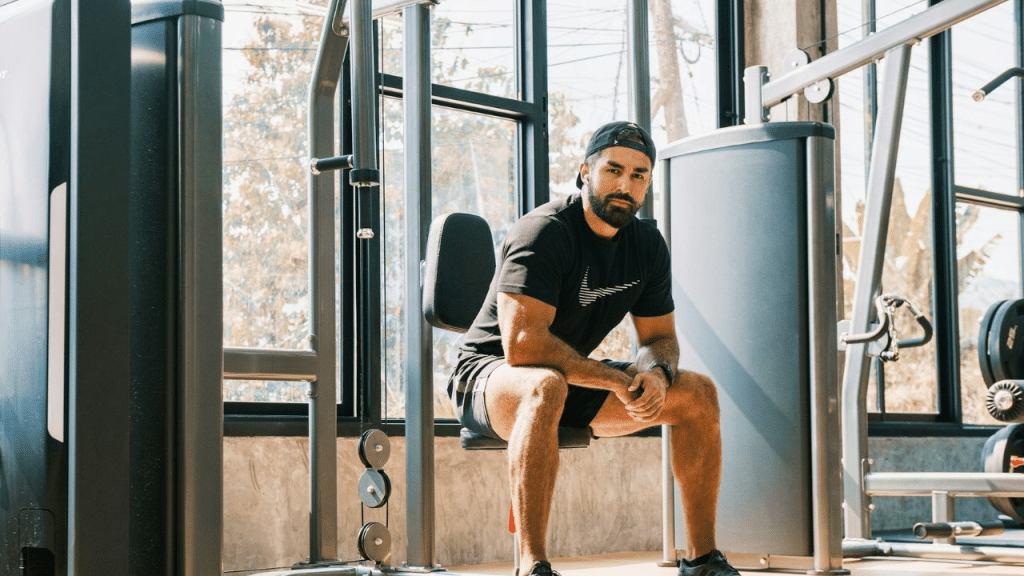 A serious bearded man in workout clothes sits on a weight machine in a sunny gym.