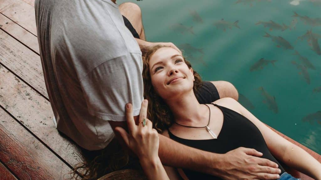 A woman lying on a man's lap by the water, smiling up at him.