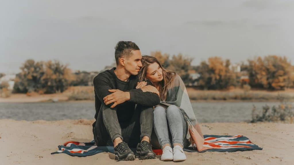A couple sitting on a blanket by the water, the woman resting her head on the man's shoulder.