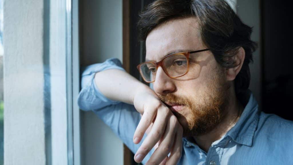 A sad-looking man with a beard and glasses stares out a window.