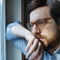 A sad-looking man with a beard and glasses stares out a window.