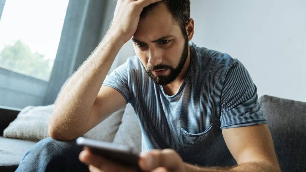 A bearded man sits, holding his head, looking stressed while viewing his phone.