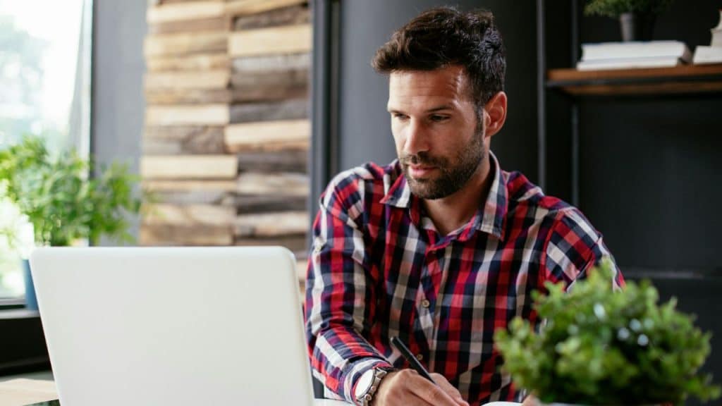 A focused man with a beard works on a laptop and writes in a notebook.