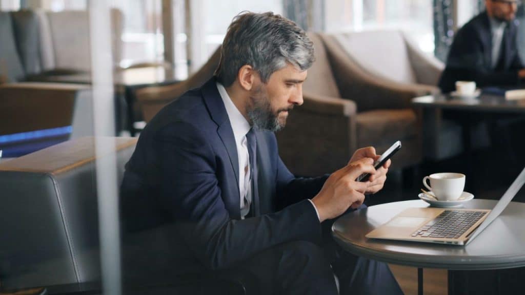 A bearded businessman in a suit uses a smartphone at a cafe table with a laptop.