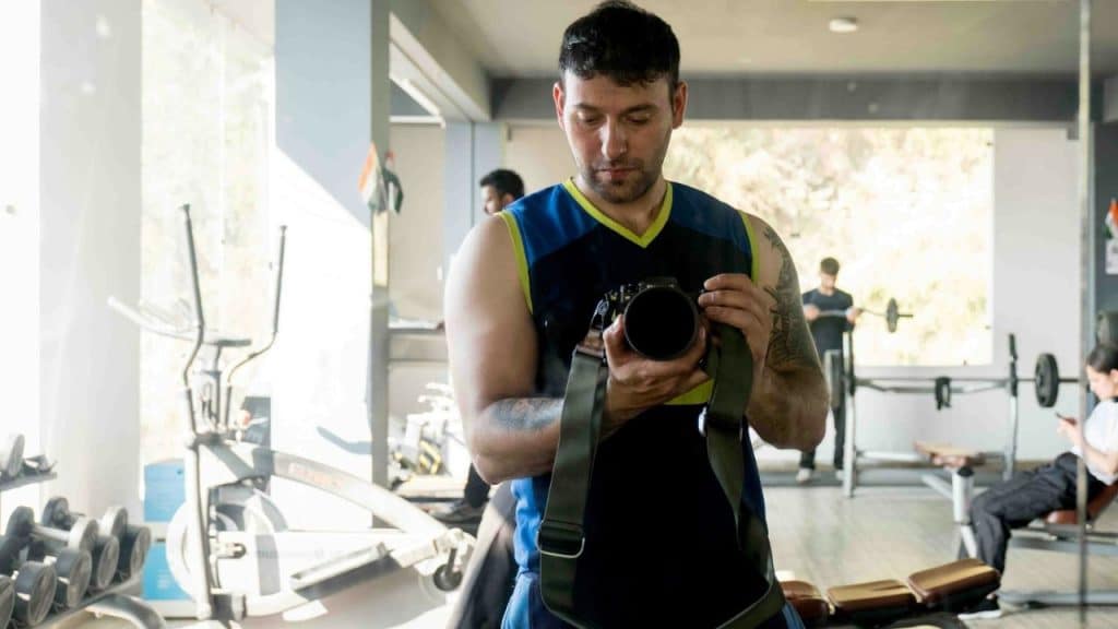 A man in a tank top takes a photo of himself in a gym mirror with a camera.