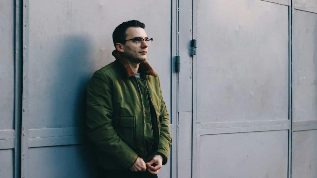 A man in glasses and a green bomber jacket stands by a gray wall.
