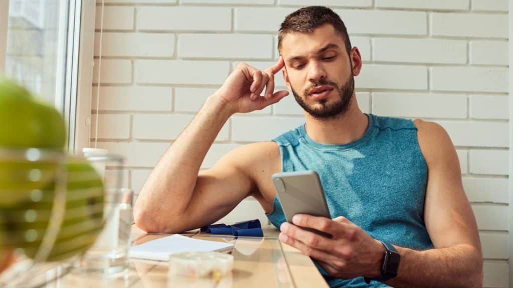 A thoughtful man in a tank top looks at his smartphone at a table indoors.