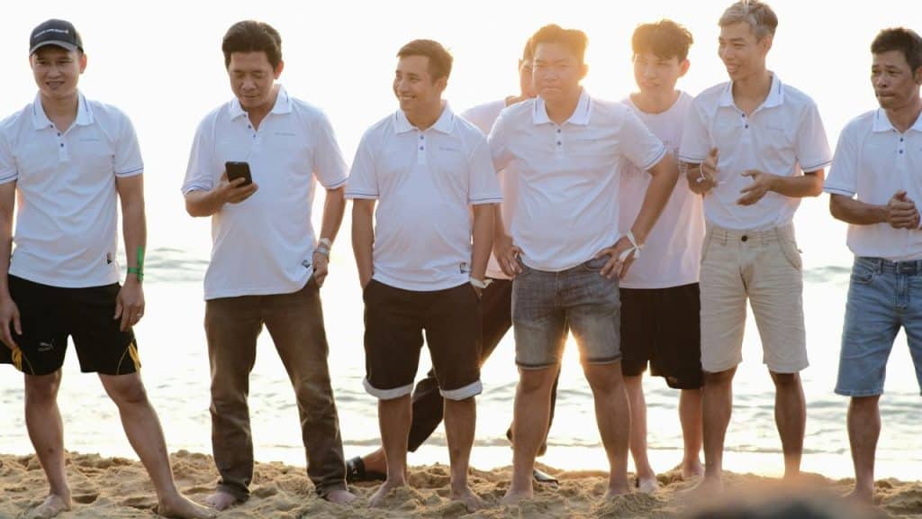 A group of eight men in white polo shirts stands barefoot on a beach at sunset.