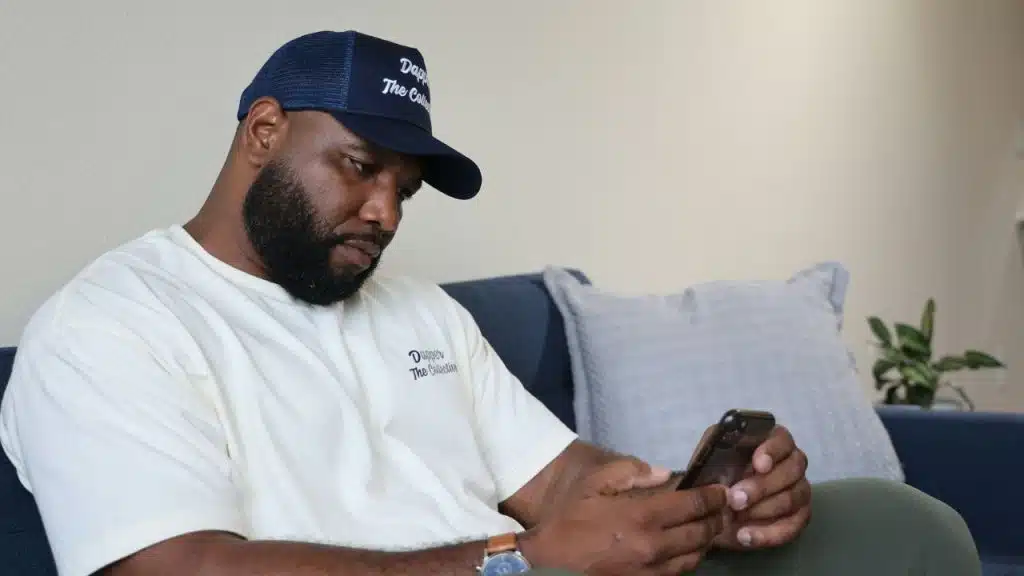 A man with a beard and a baseball cap sits on a couch using his phone.