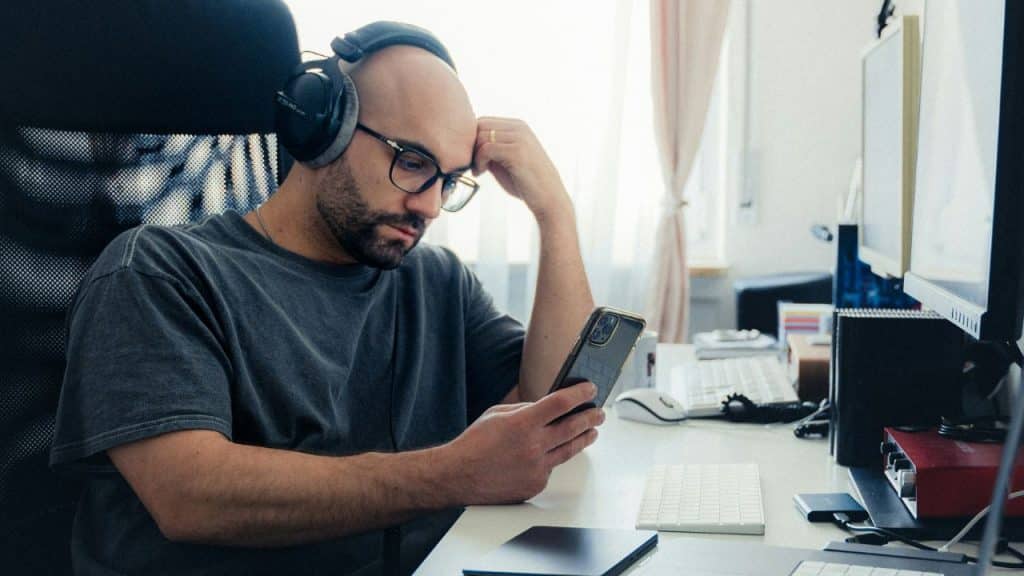 A bald man with glasses and headphones looks intently at his smartphone at a desk.