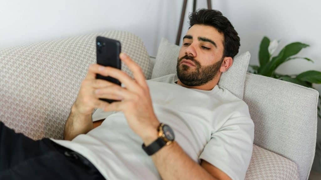 A serious man lies on a couch, propped up with pillows, looking at his smartphone.