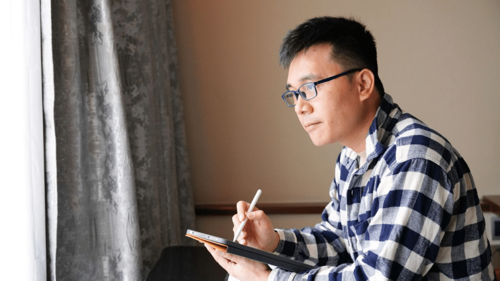 A focused man wearing glasses and a plaid shirt sketches on a tablet near a window.