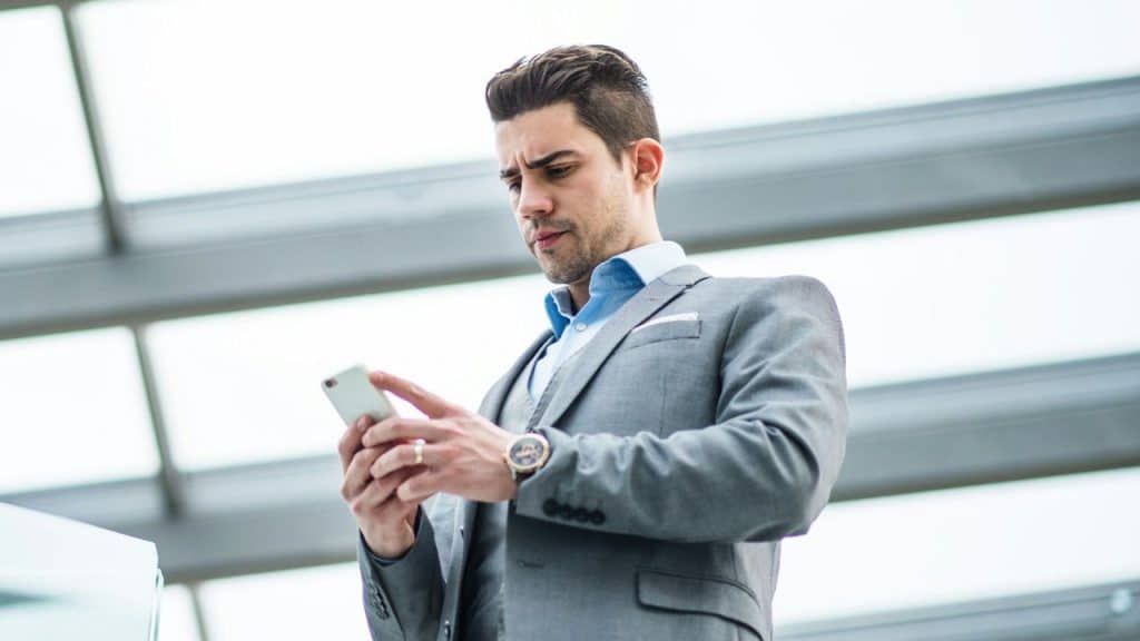 A focused man in a gray suit looks down at the smartphone he holds in both hands.