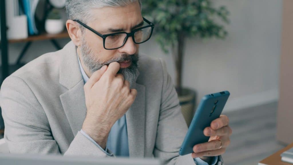 A thoughtful, gray-bearded man in glasses looks at his phone, biting his nail.
