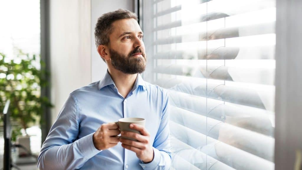 A pensive, bearded man in a blue shirt holds a mug, looking out a window.