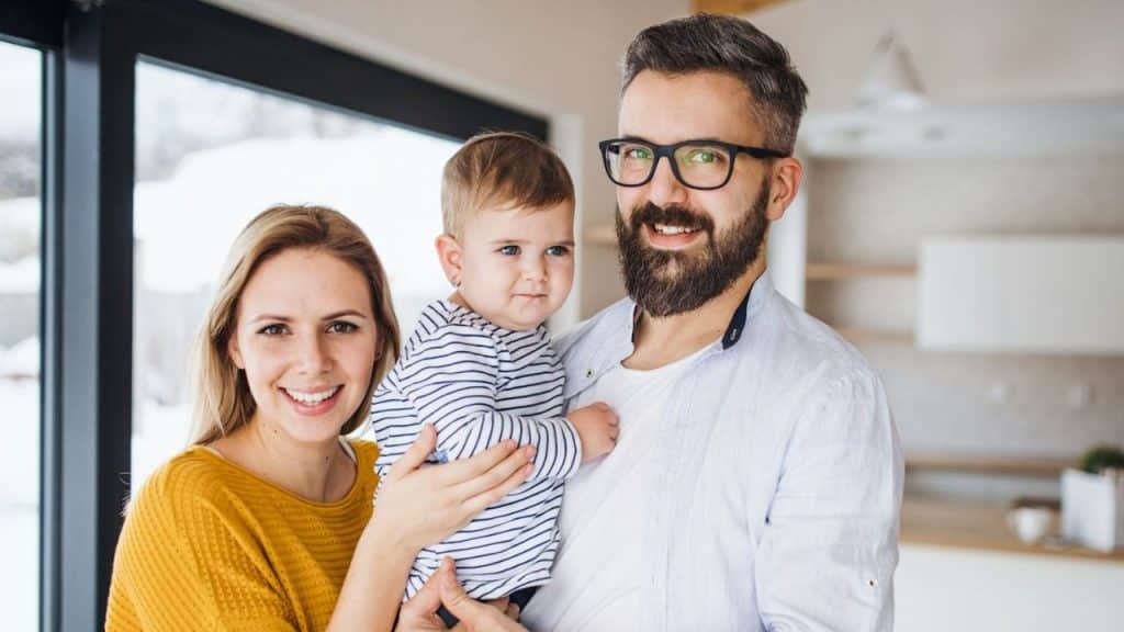 A smiling couple and their baby are standing together in a bright, modern room.