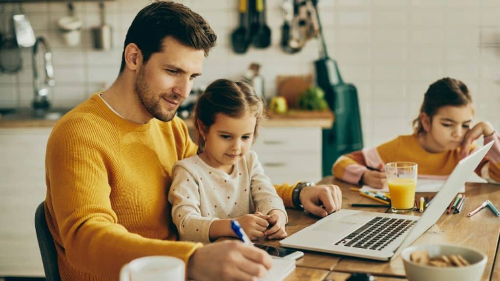 A father works on a laptop at a kitchen table while his two daughters sit nearby.