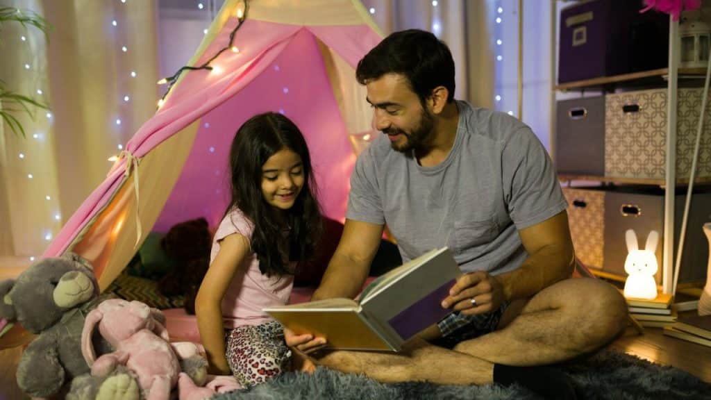 A happy father reads a book to his young daughter inside a fairy-light-lit indoor tent.