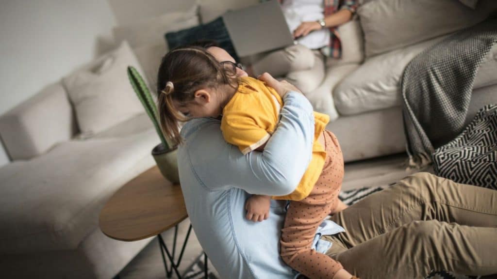 A man sits on the floor holding a young girl in an orange shirt close to his chest.