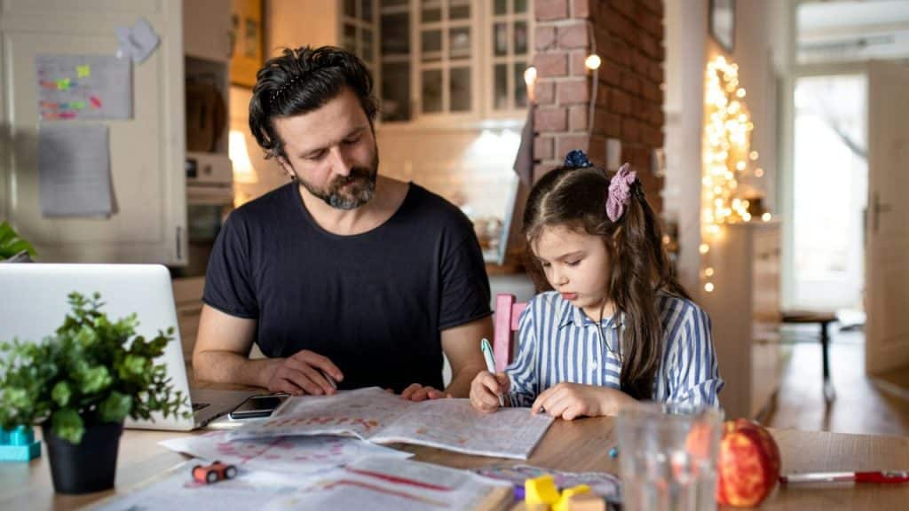 A serious man watches as a young girl draws or writes on paper at a kitchen table.
