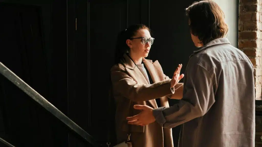 A woman in glasses and a tan coat holds her hand out toward a man.