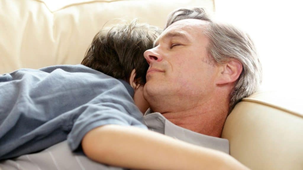 An older man rests on a sofa, embracing a child whose face is hidden from view.