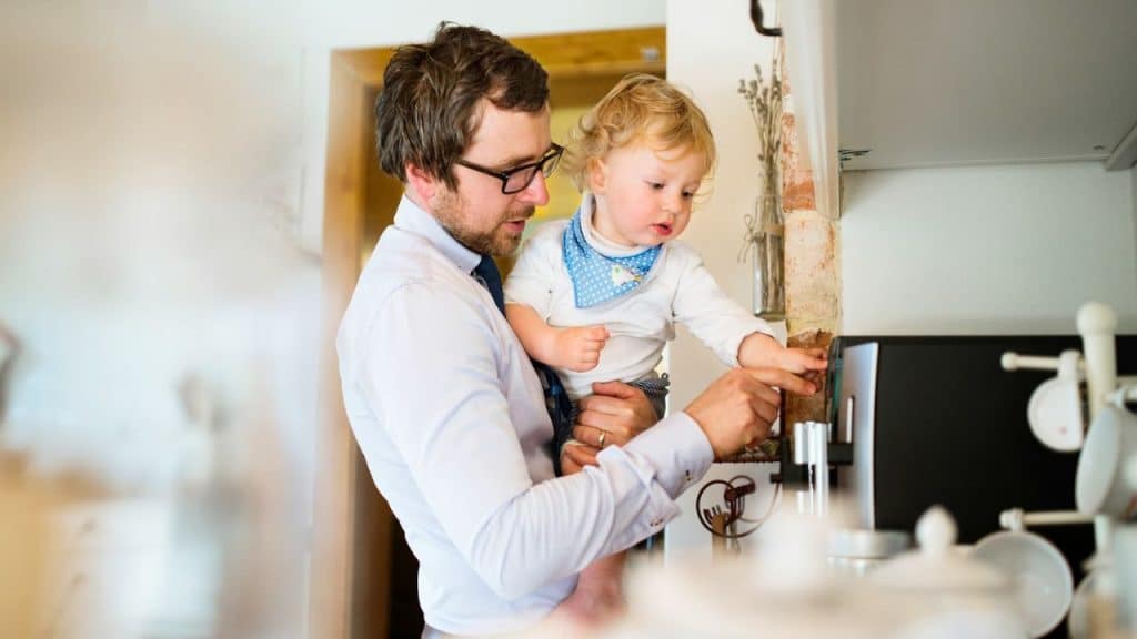 A father in a dress shirt holds a toddler who is pressing a button on a coffee machine.