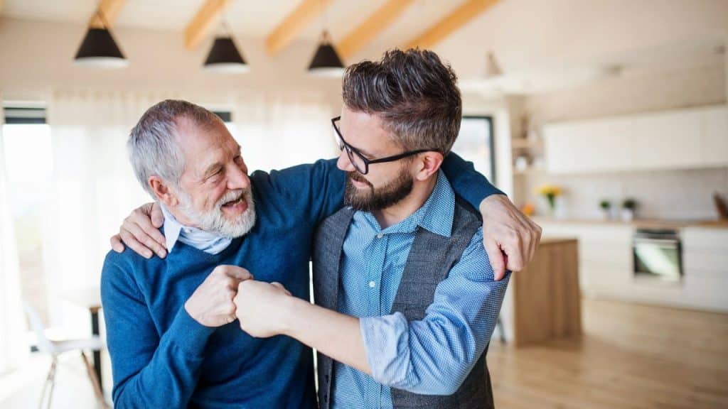 An older man and a younger man hug and fist-bump while standing in a bright kitchen.