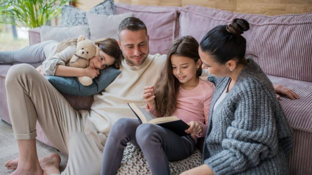 A father, mother, and two daughters sit together on the floor reading a book in a living room.