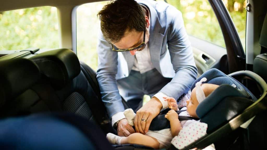 A man in a suit carefully buckles a baby into a car seat in the back of a car.