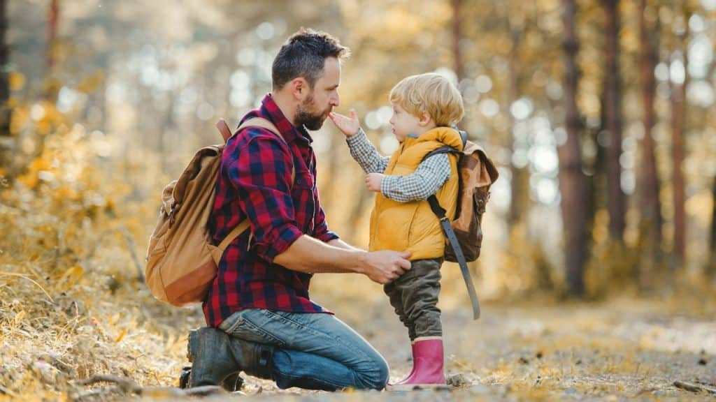 A father kneels on a forest path as his young son touches his beard.