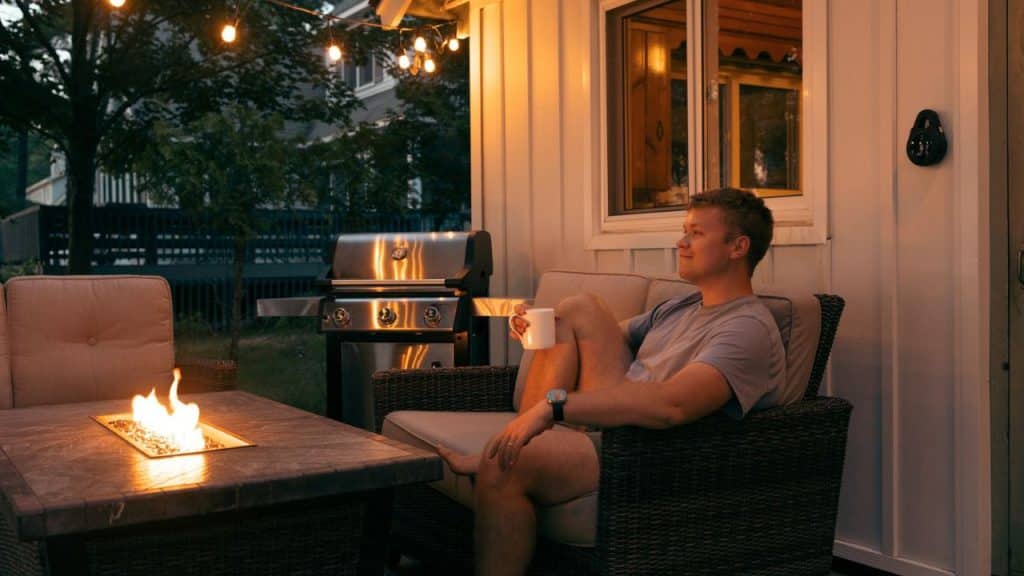 A man sits in an outdoor chair with a mug near a fire pit and grill at twilight.